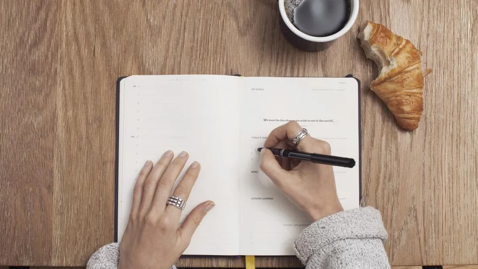 Mão escrevendo num caderno aberto sobre mesa de madeira, com café ao lado
